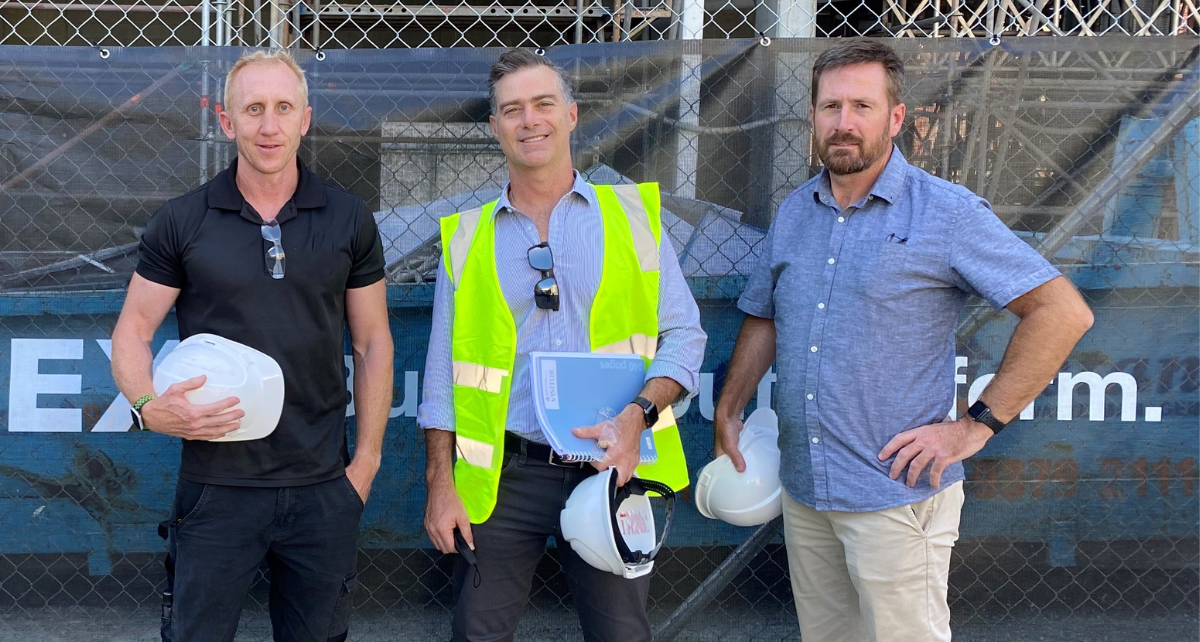 Three men dressed in construction attire smiling in front of a building site.