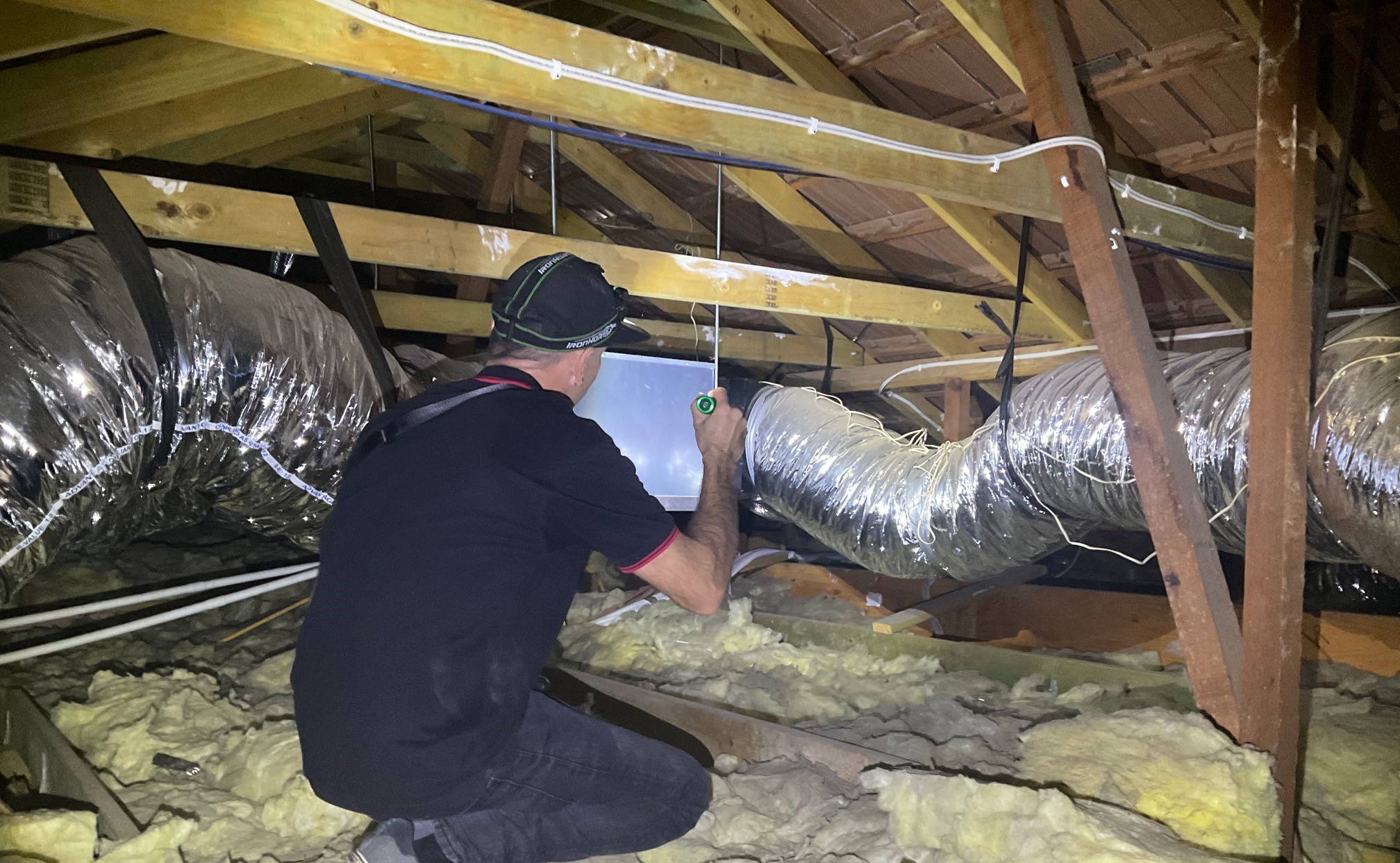 Building inspector conducting a roof space inspection with flashlight, assessing ductwork and insulation in a new home in Busselton.
